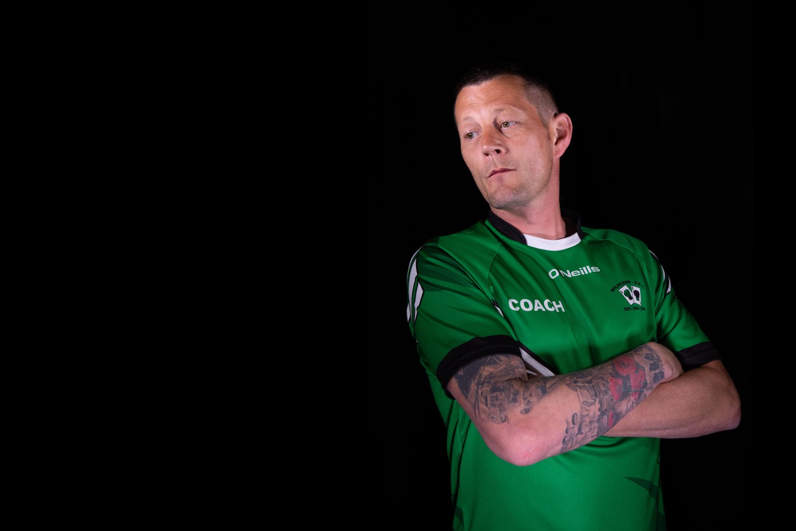 A man with tattoos on one arm, wearing a  green sports top stands with crossed arms and looks to one side.