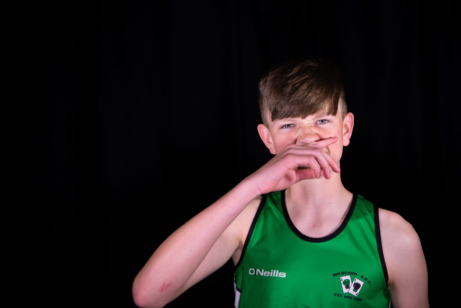 A young person with dark hair, wearing a green sports vest wipes their nose with their hand.