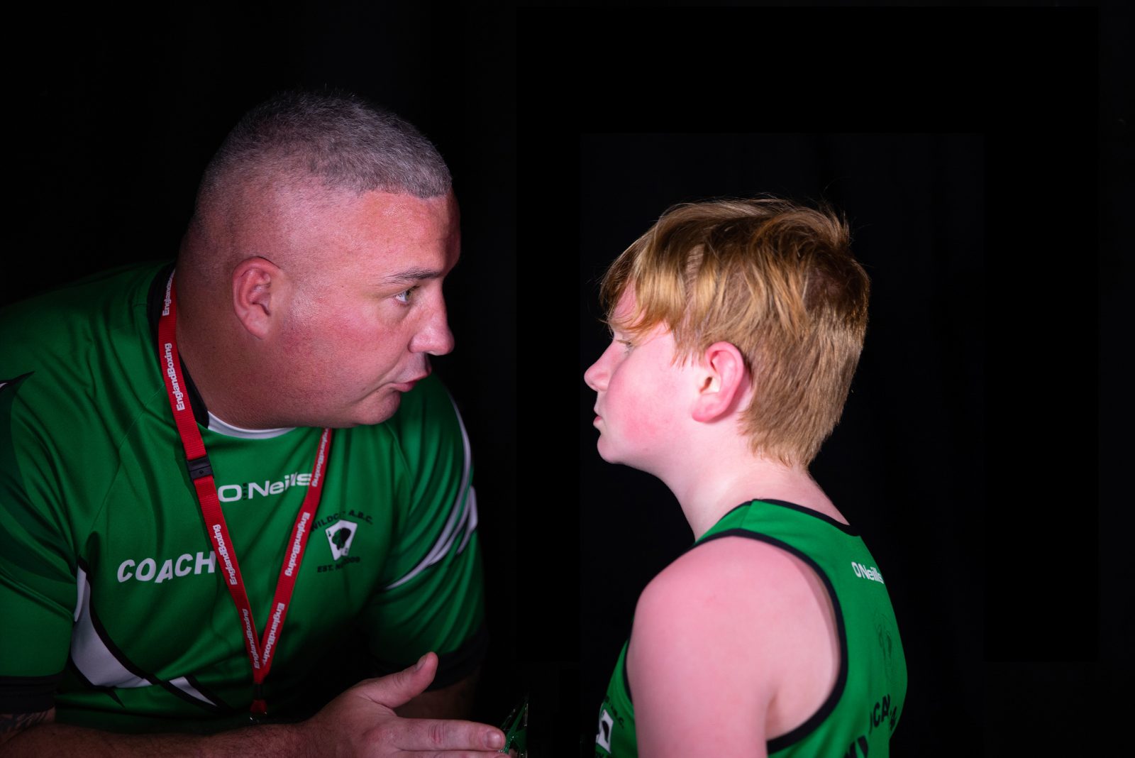 A man wearing a green sports top and a red lanyard is talking to a young person in a green sports vest. The man is gesturing with their hand. The young person is red faced,