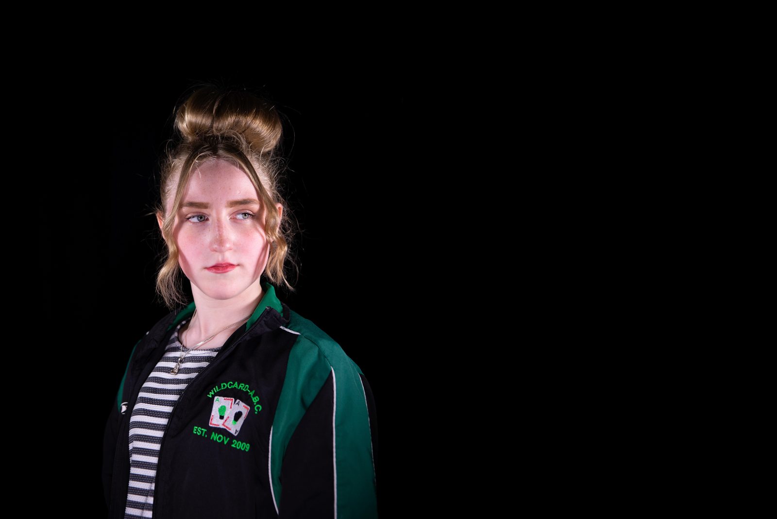 A young woman wearing a striped top and a black and green sports jacket stands a looks to one side.