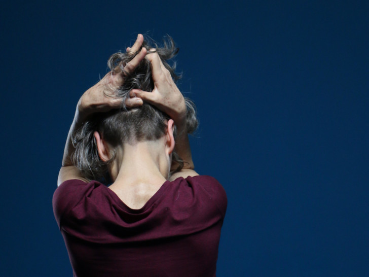 A person wearing a maroon short sleeved t-shirt and with short cropped hair at the back and longer hair on top runs their hands through the top of their hair. They are facing away from the camera against a navy blue background.