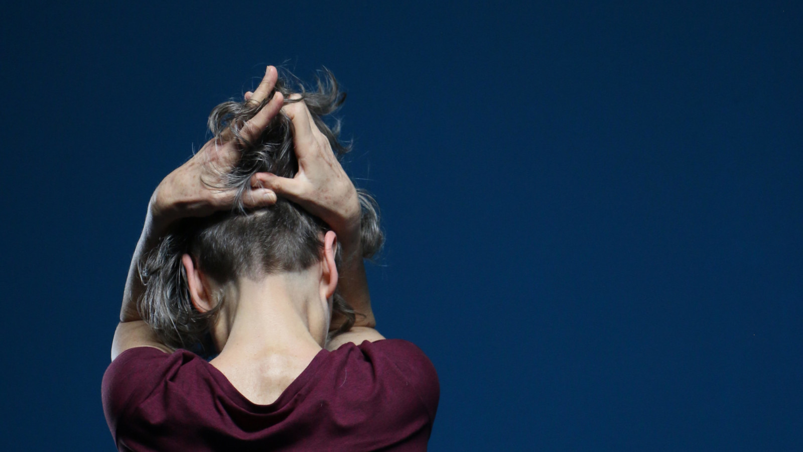 A person wearing a maroon short sleeved t-shirt and with short cropped hair at the back and longer hair on top runs their hands through the top of their hair. They are facing away from the camera against a navy blue background.