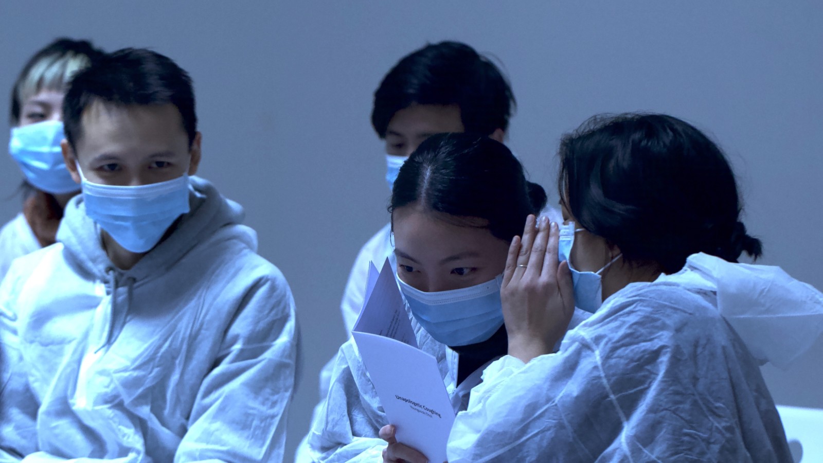 A group of East Asian audience members wear face masks and white overalls. A person whispers into the ear of another person who is holding a performance text. Other audience members watch.
