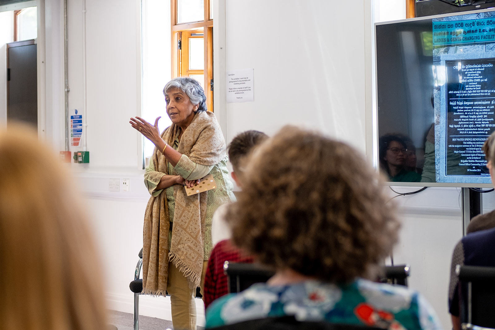 Radha D'Souza is giving a talk inside in front of a screen, she his talking with her hands and wears all neutral colours, with a patterned scarf.