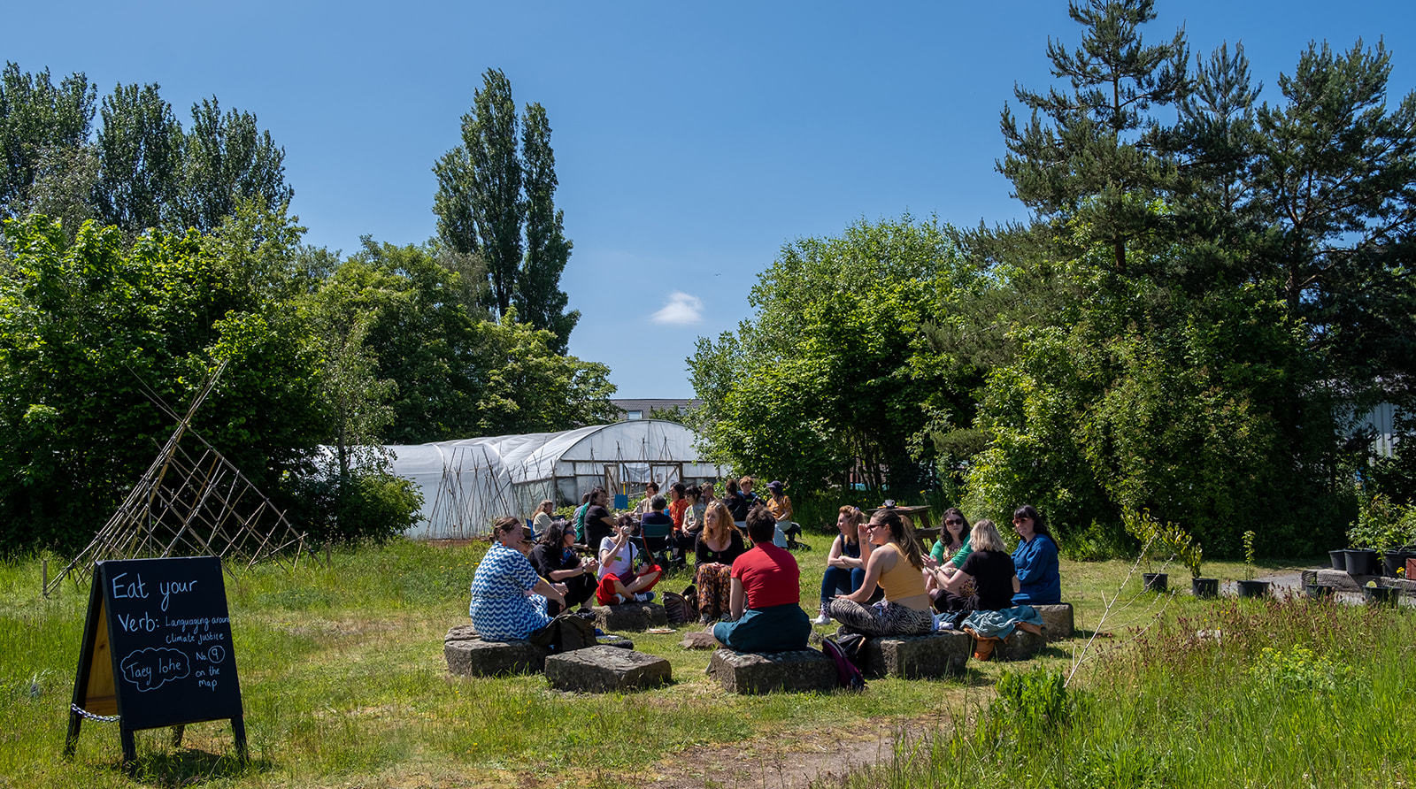 People are sat in the community garden at Incredible Edible Knowsley, it is sunny and there is a sign that reads 'Eat your Verb'