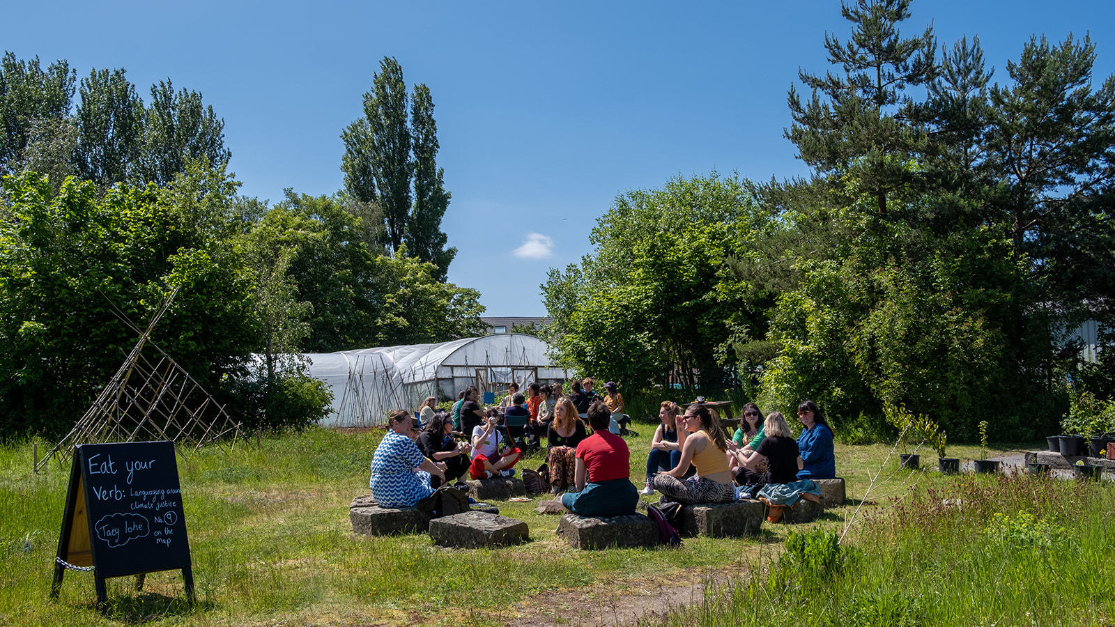 People are sat in the community garden at Incredible Edible Knowsley, it is sunny and there is a sign that reads 'Eat your Verb'