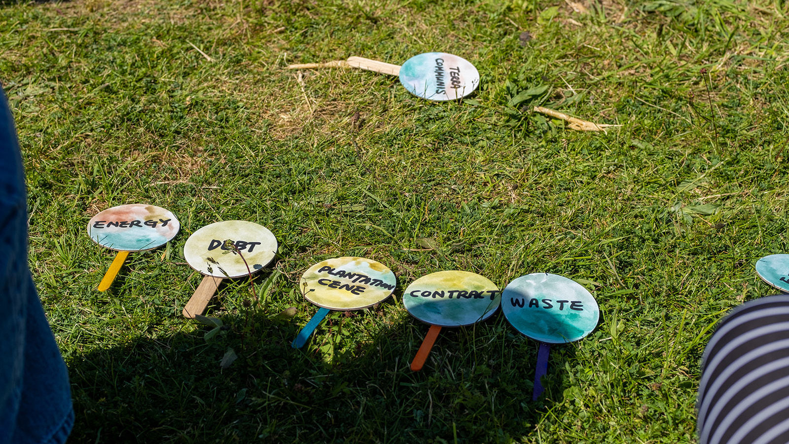 Mini signs are laid out on the grass that read 'Terra Communis', 'Energy', 'Debt', 'Plantation cane', 'Contract', and 'Waste'.