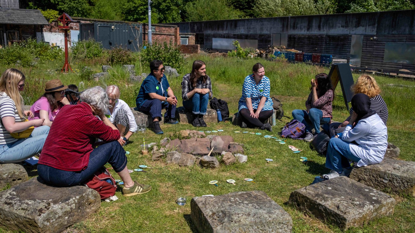 A group of people sit on stones in a circle, outdoors on a sunny day. They are taking park in Taey Iohe's workshop 'East your Verb'.