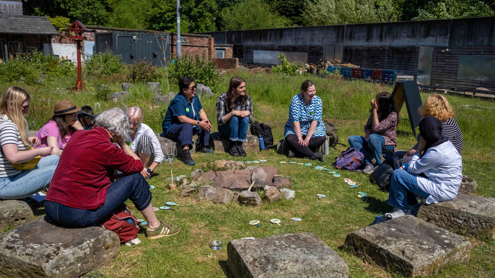 A group of people sit on stones in a circle, outdoors on a sunny day. They are taking park in Taey Iohe's workshop 'East your Verb'.