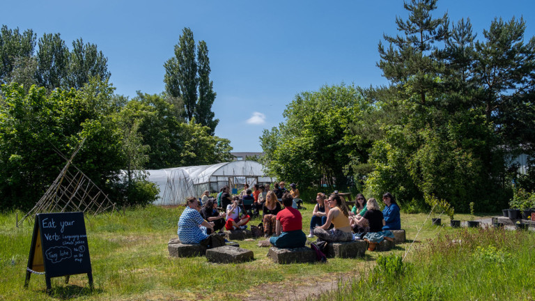 People are sat in the community garden at Incredible Edible Knowsley, it is sunny and there is a sign that reads 'Eat your Verb'