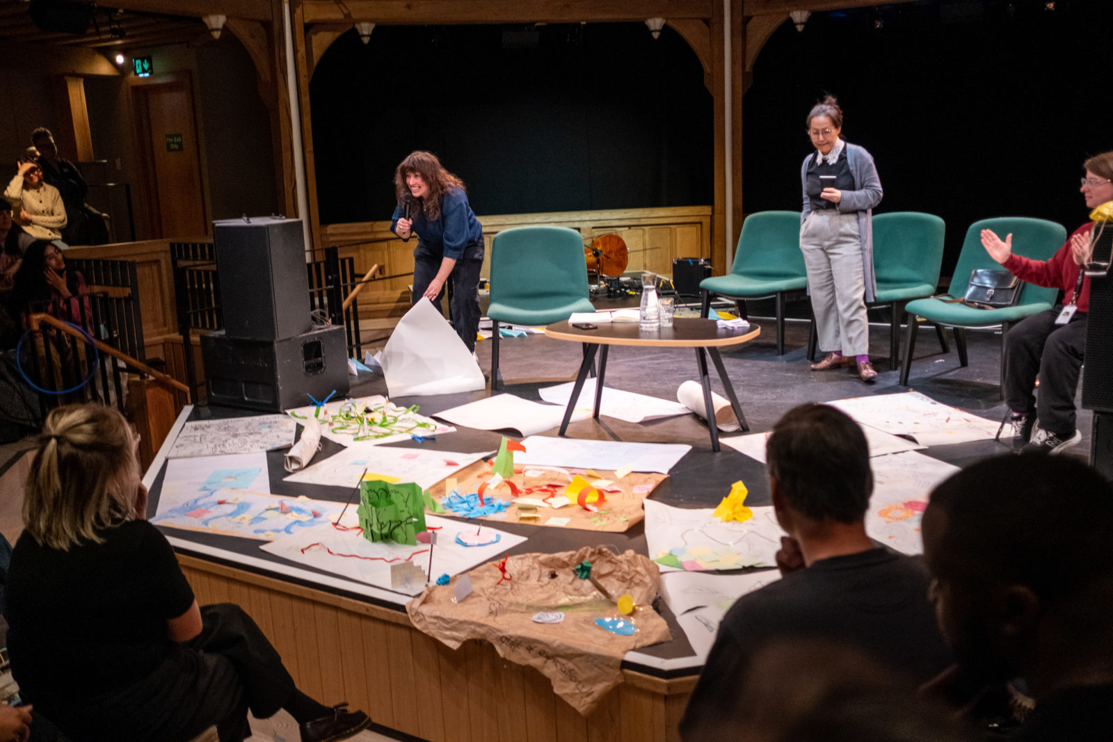 Three women are on a stage. One with long brown hair holds a microphone in one hand and holds up a large sheets of decorated paper in the other. There are more sheets of decorated paper lying on the stage.