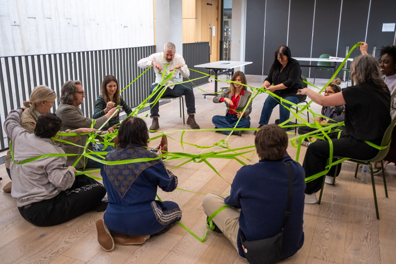 A group of people sit in a circle on the floor and on chairs. Wrapped around them, connecting the group is a bright green web of tape.