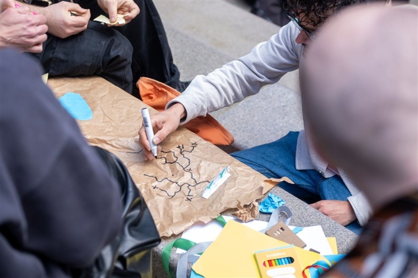Someone draws a map on a large piece of brown paper while other sit around watching and folding pieces of card.