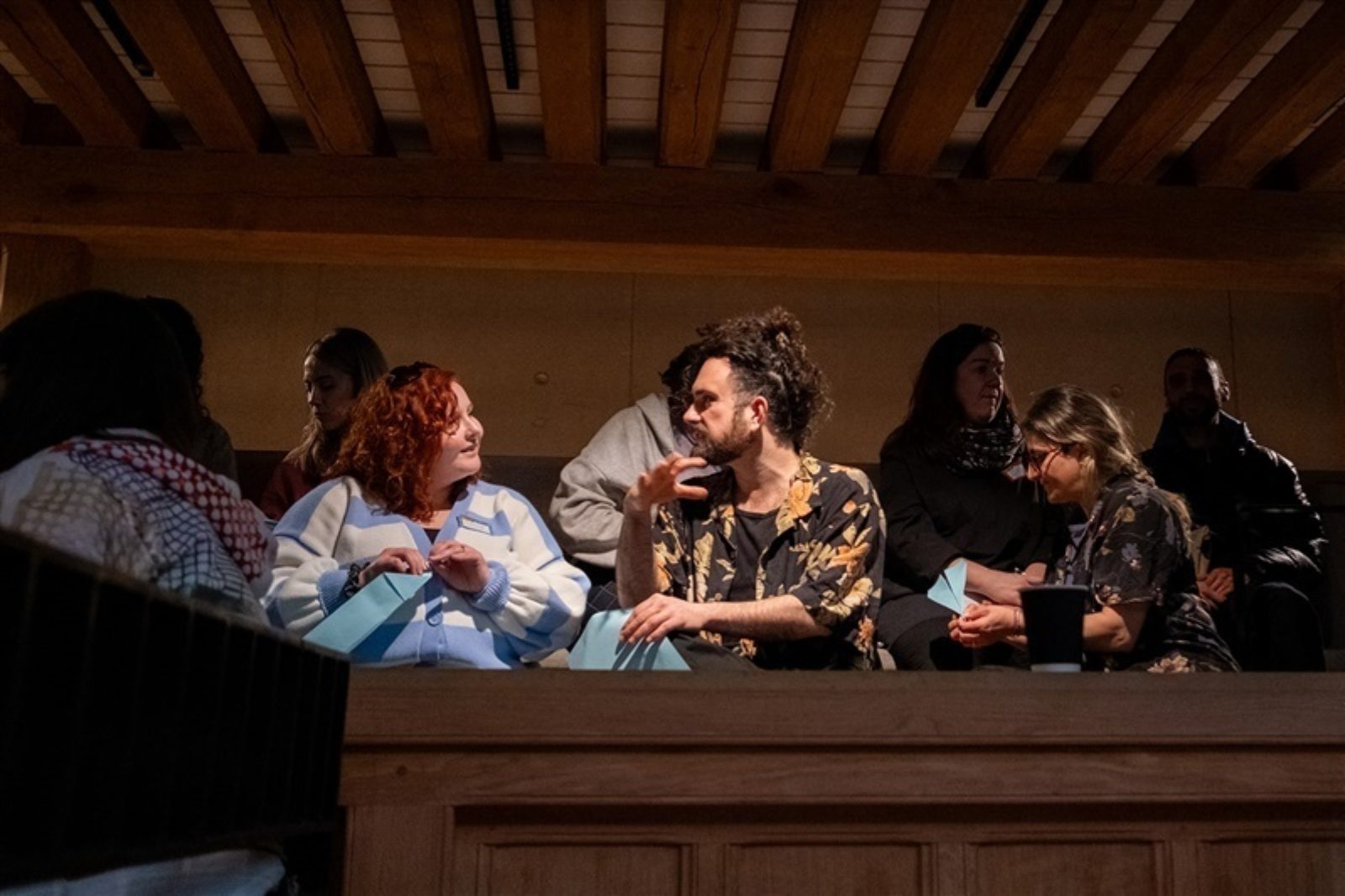 Some people sit in a low-lit auditorium. Two of the audience members are in conversation, one is folding a paper aeroplane.
