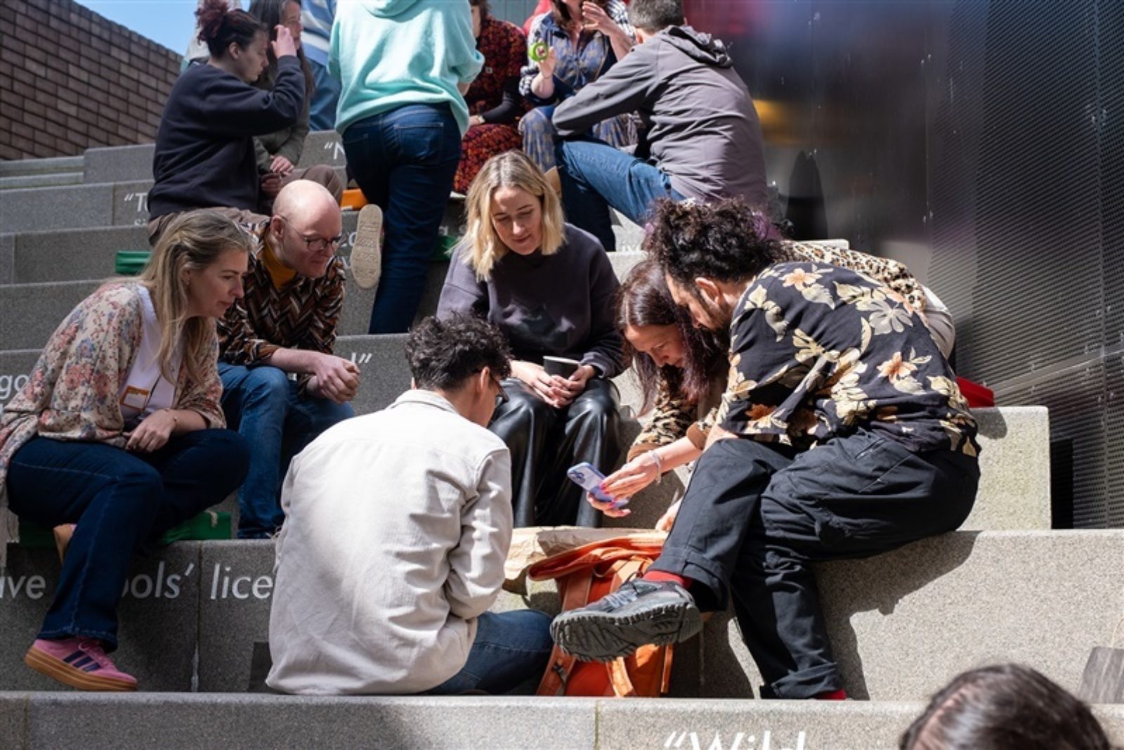 A small group of people sit on large concrete steps. They are looking at and taking photographs of something on the ground.
