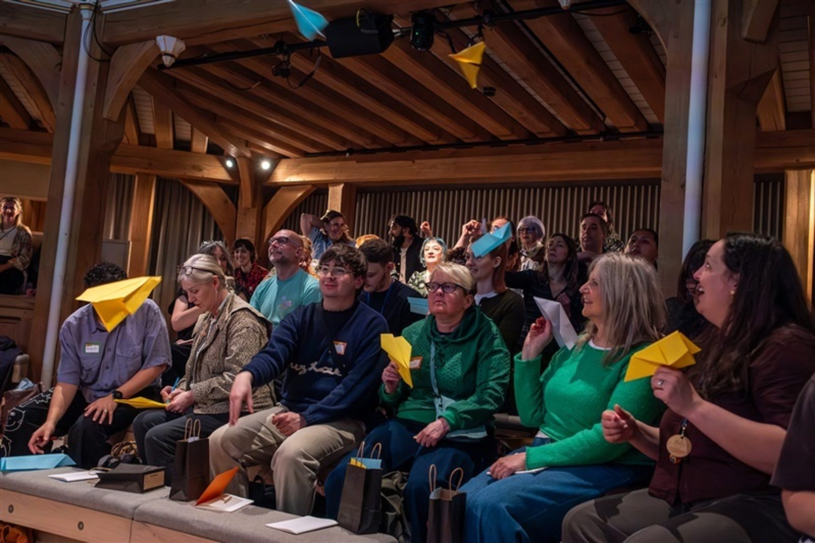 An audience sit is an auditorium in low lighting. Colourful paper aeroplanes fly in front of them towards the stage.