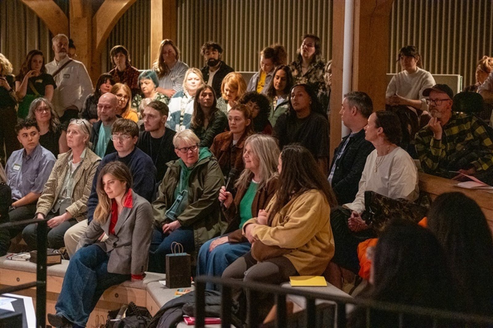 An audience of people sit in an auditorium. One of the audience members holds a microphone and is speaking while the audience listen.