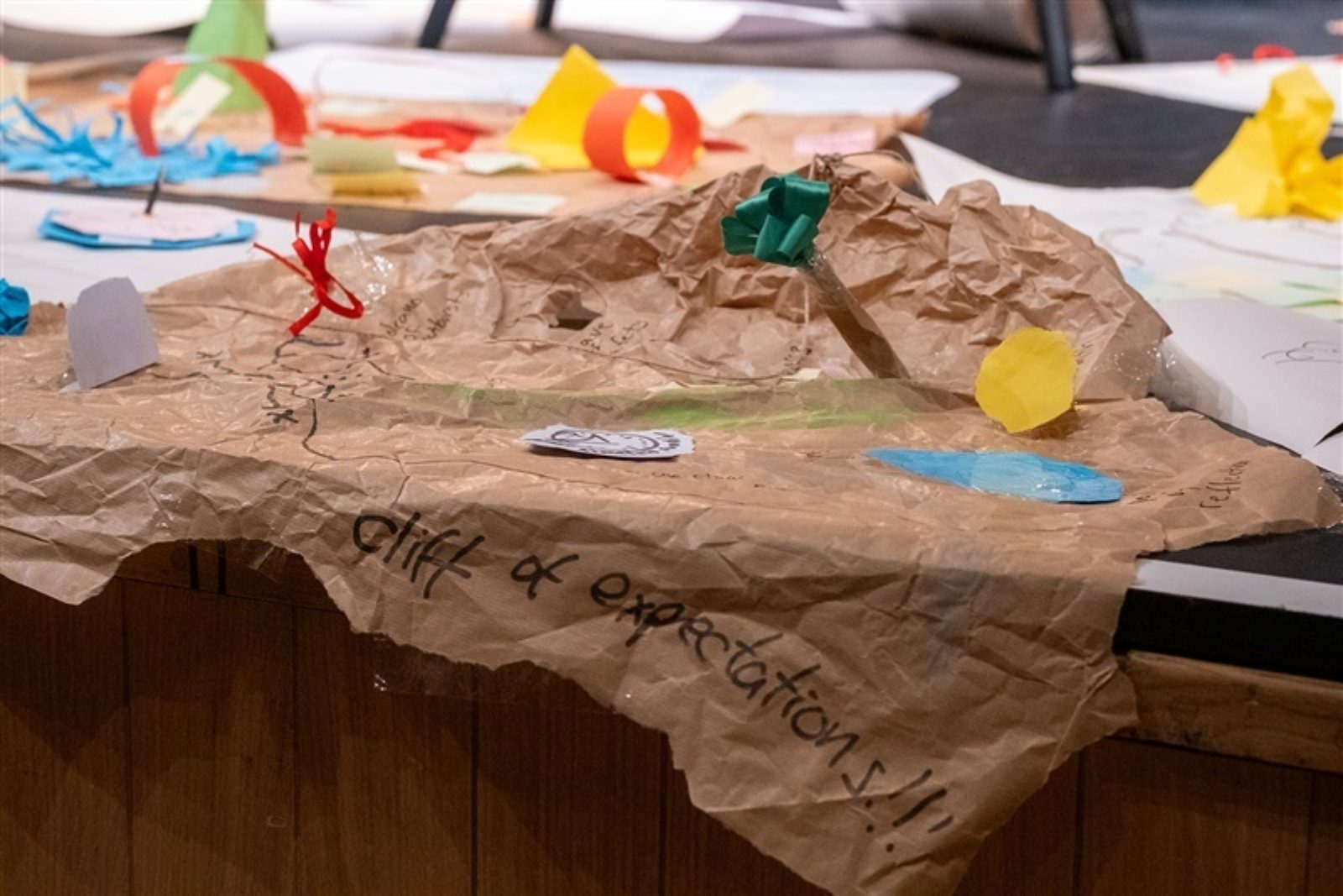 Some large pieces of paper lie on a stage. One large, crinkled piece of brown paper has a paper tree standing on it and a section hanging over the stage that reads 'cliff of expectations'