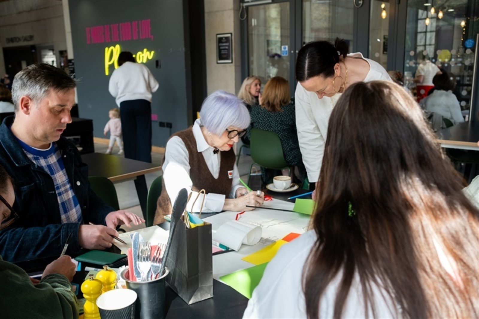 A group of people, including Chrissie Tiller are gathered at a table, writing on and adding colourful paper to a large sheet of paper.
