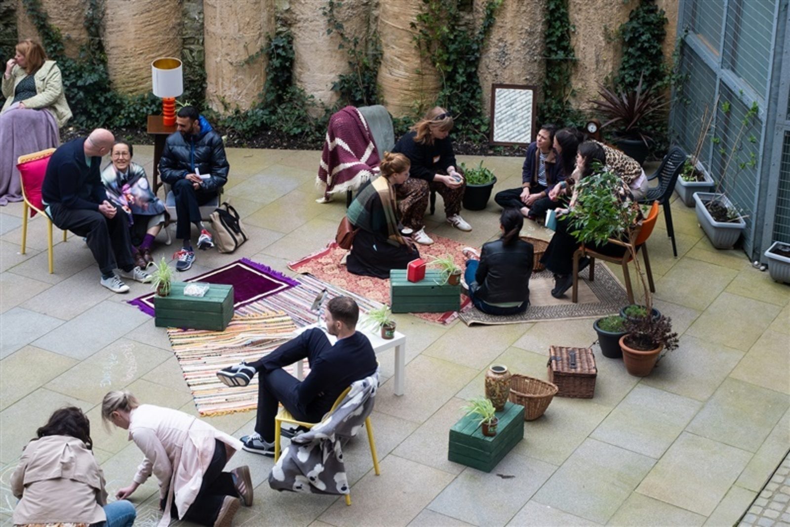 In a courtyard with rugs, seating, coffee tables and plants people sit in small groups talking, writing on the floor with chalk and laughing.