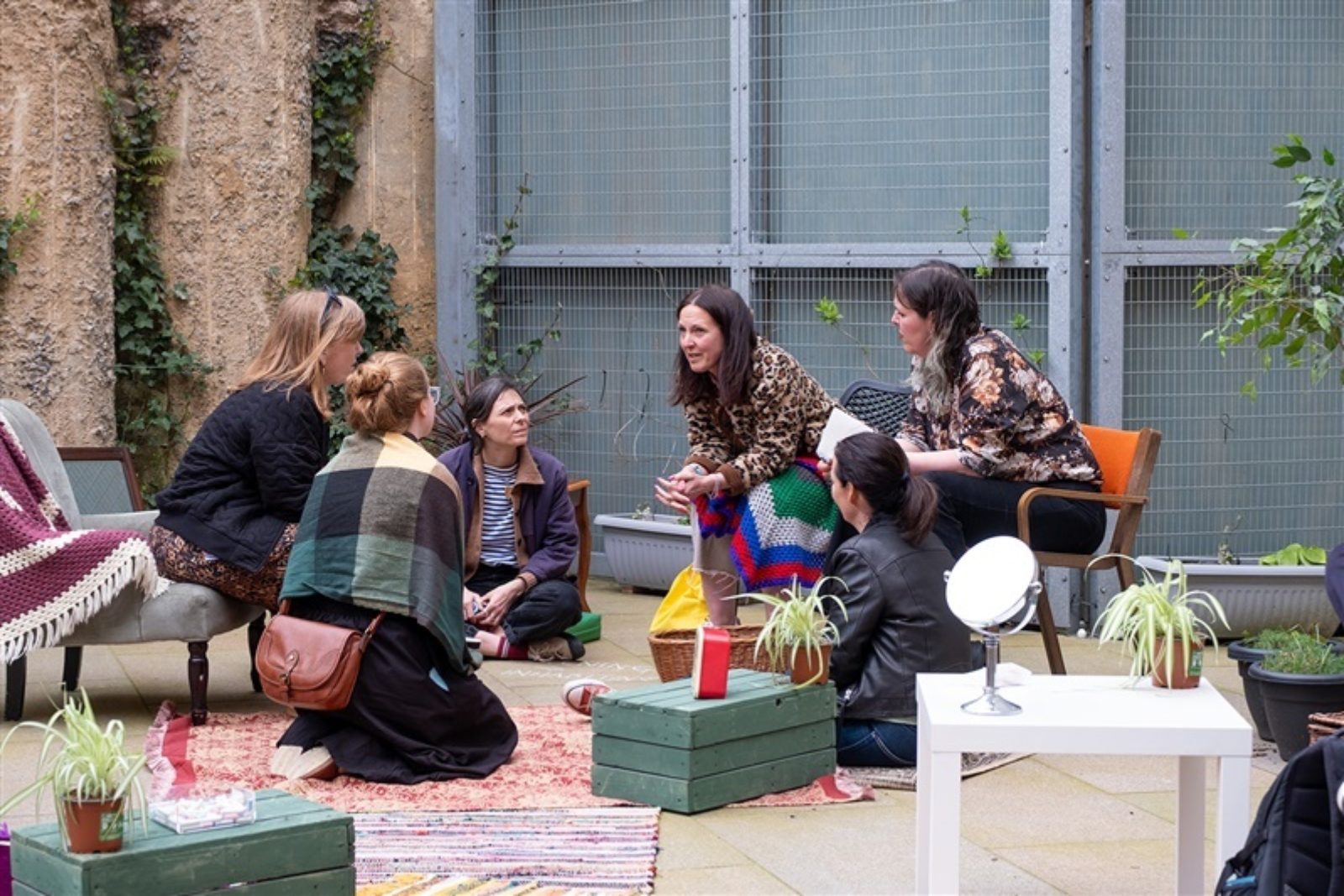 A group of people sit in a circle in conversation. They sit on rugs, chairs and cushions surrounded by other decorative household items