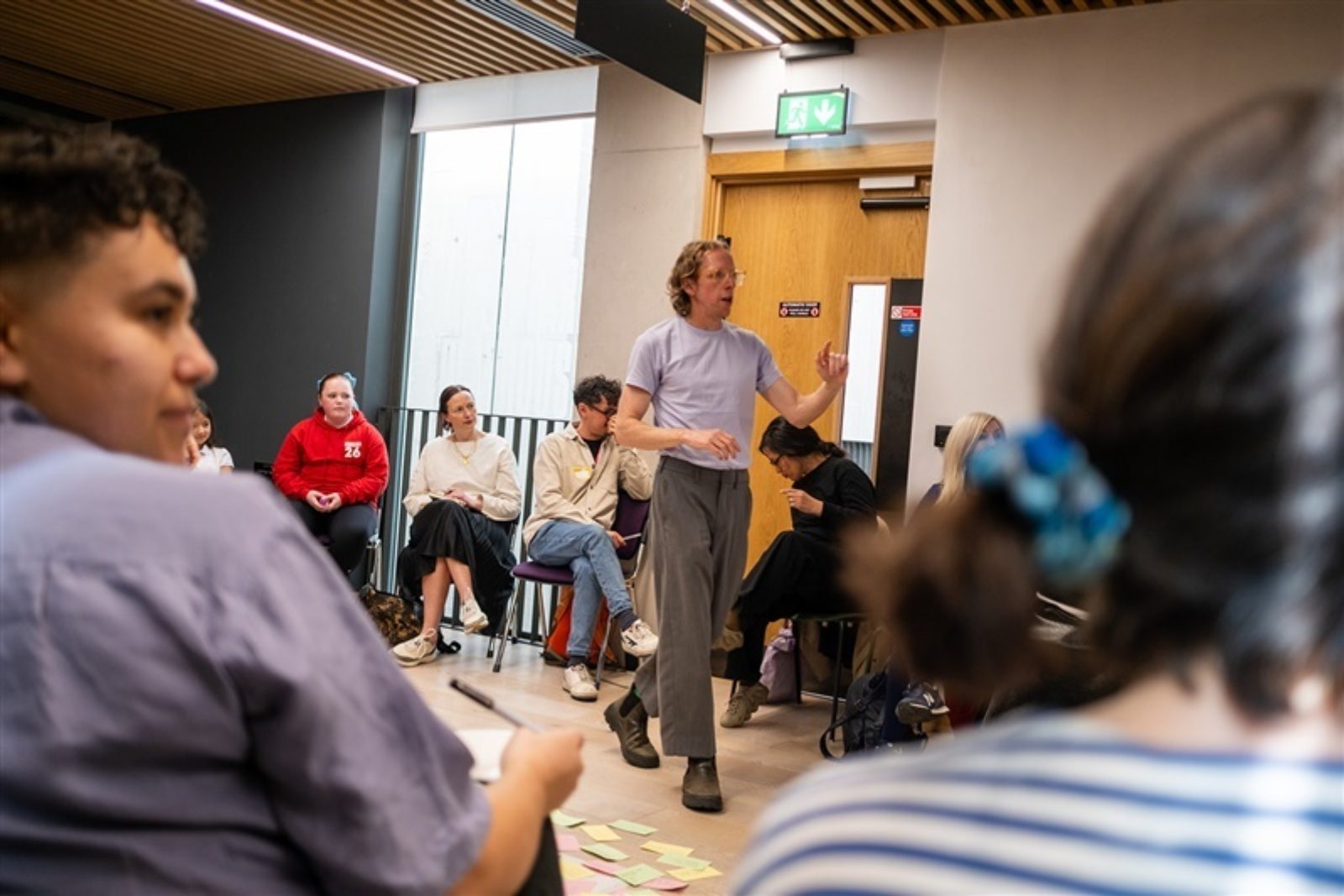 A man stands in the middle of a circle of people seated in a light room. He is talking while the audience look at him and off screen. The audience is made of children and adults.