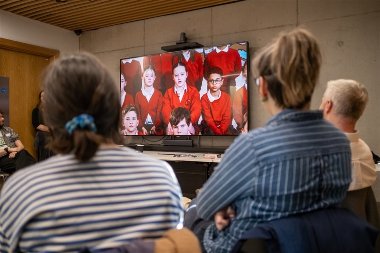 A large television screen shows a group of school children in red cardigans sat in a row. Some adults sit watching the screen.