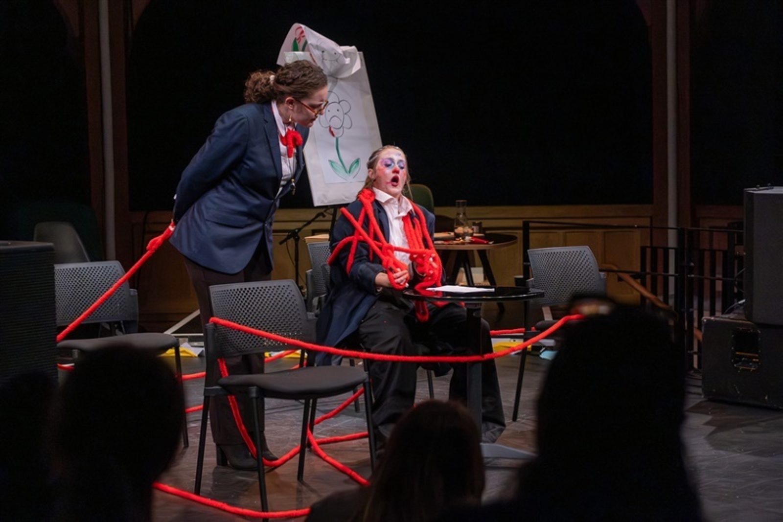 Two performers stand on a stage wearing dark blue uniforms. One wears clown like makeup and wrapping around them and the stage is a long red string which is held in the hands of the other person.