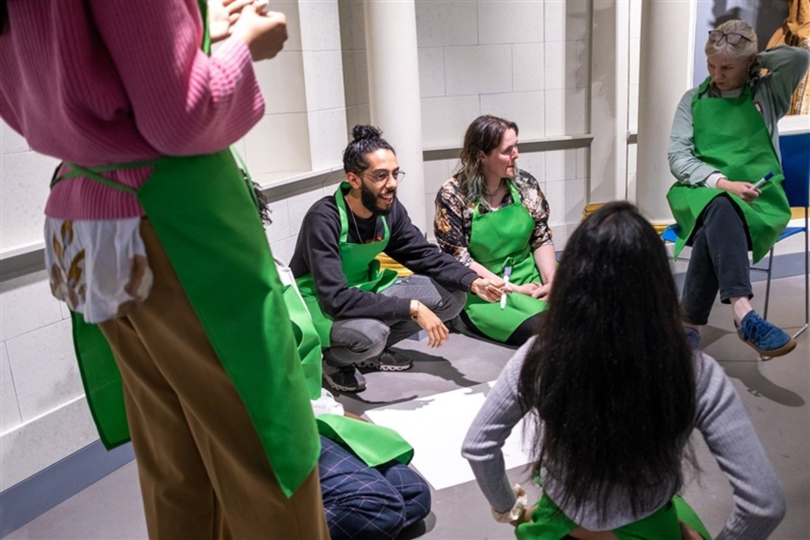 A group of people are gathered around a large piece of paper.. They sit, kneel and stand while in conversation. Each person is wearing a green apron.