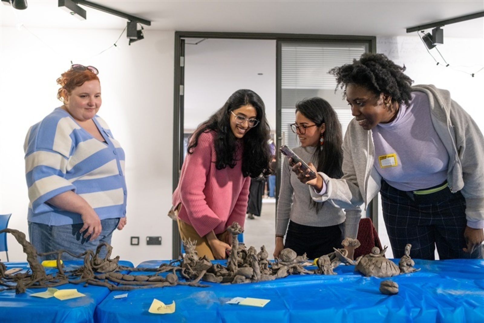 Four people stand in front of a table covered in blue tarpaulin and clay models. They are taking photos, looking at the models and smiling.