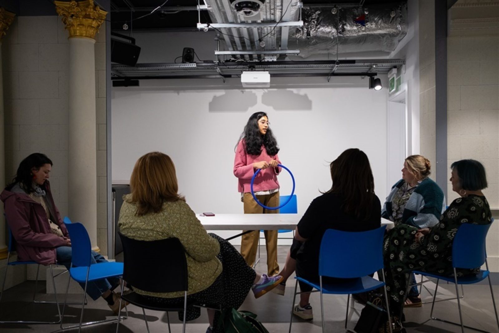 A group of five people sit around a trestle table. In front of them a woman stands holding a small hula hoop and talking to the group.