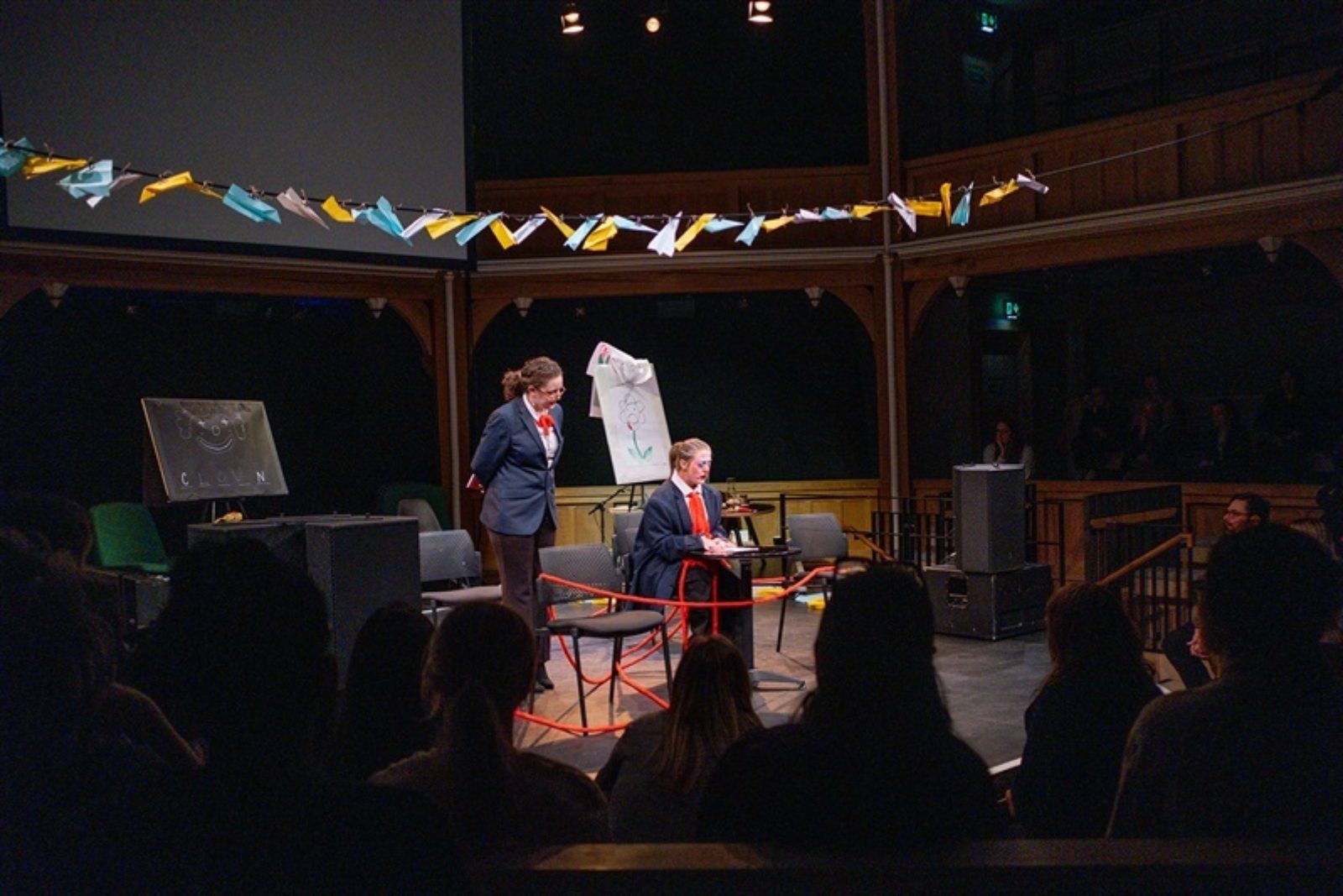 A string filled with colourful paper aeroplanes hangs over a stage. On the stage two performers stand wearing dark blue uniforms, wrapping around them and the stage is a long red string.
