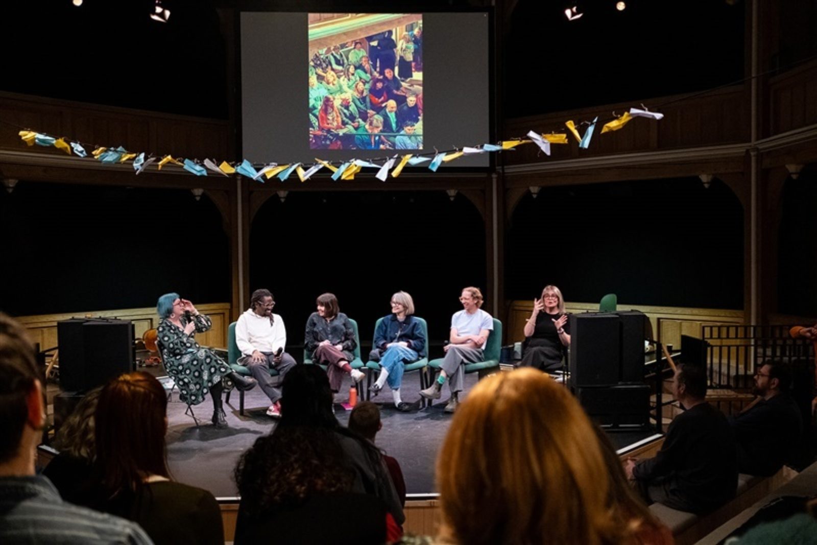 Five people sit on stage in a line having a conversation. Hanging above them is a string filled with colourful paper aeroplanes. Alongside them another person provides BSL interpretation.