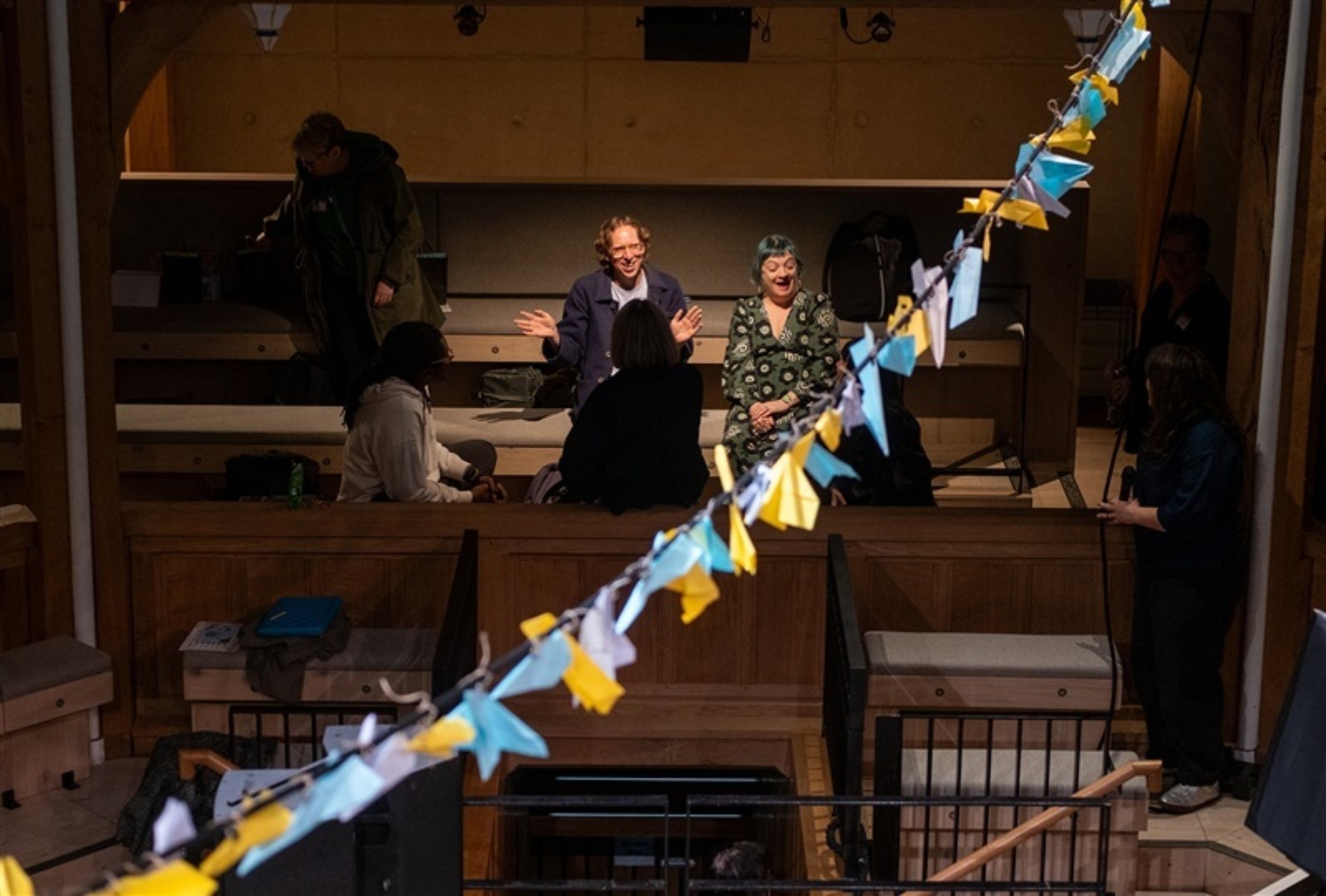 A string full of colourful paper aeroplanes hangs across a stage. In a low lit auditorium a small group of people sit in conversation smiling.