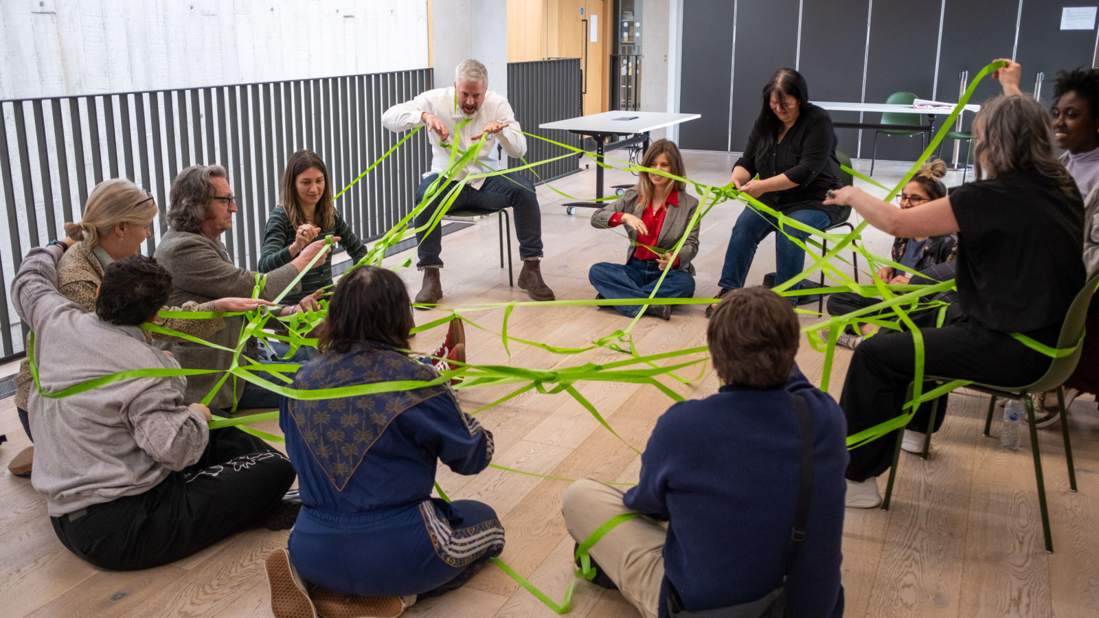 A group of people sit in a circle on the floor and on chairs. Wrapped around them, connecting the group is a bright green web of tape.