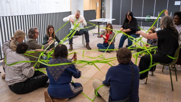 A group of people sit in a circle on the floor and on chairs. Wrapped around them, connecting the group is a bright green web of tape.