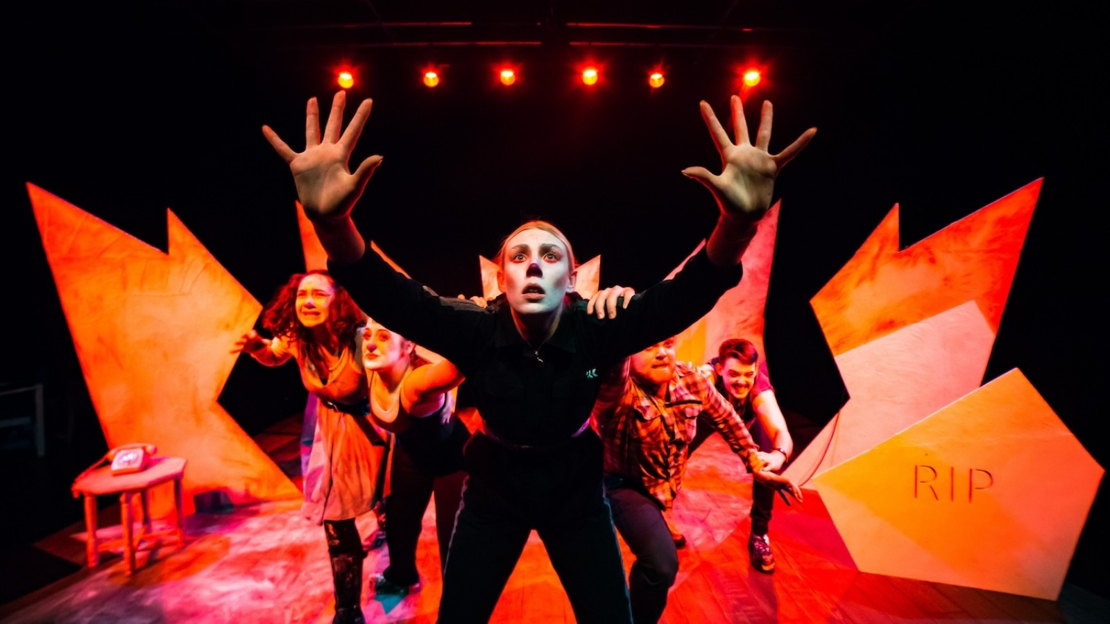Performers in white face paint on a stage, with shocked faces and hands reaching towards the camera. Behind them a jagged paper set and orange lights/