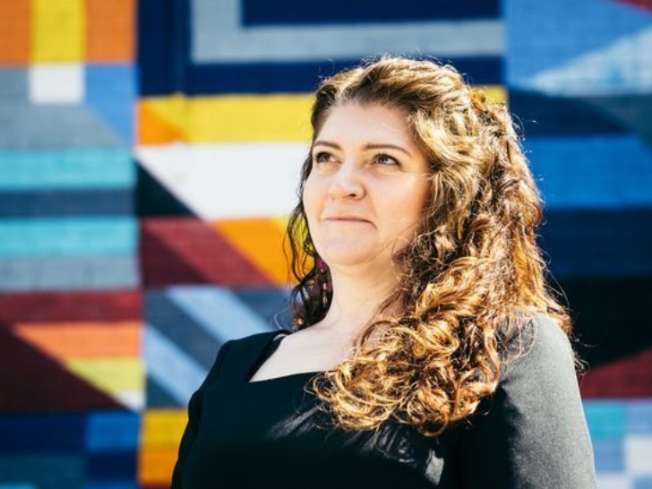 A woman with curly brown hair wearing a black dress. She is smiling and stood in front of a colourful background.