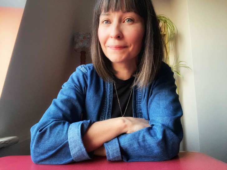 Gina Tsang is photographed with her arms folded on the table in front of her, she has medium length, straight dark hair with a fringe and wears a denim shirt.