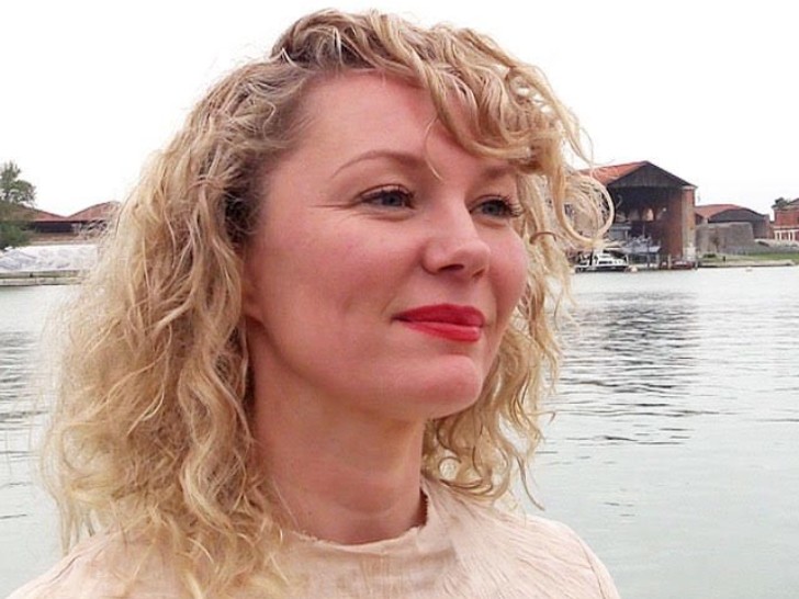 Jesse is photographed looking past the camera smiling in front of a lake. She has blonde curly hair and wears red lipstick.