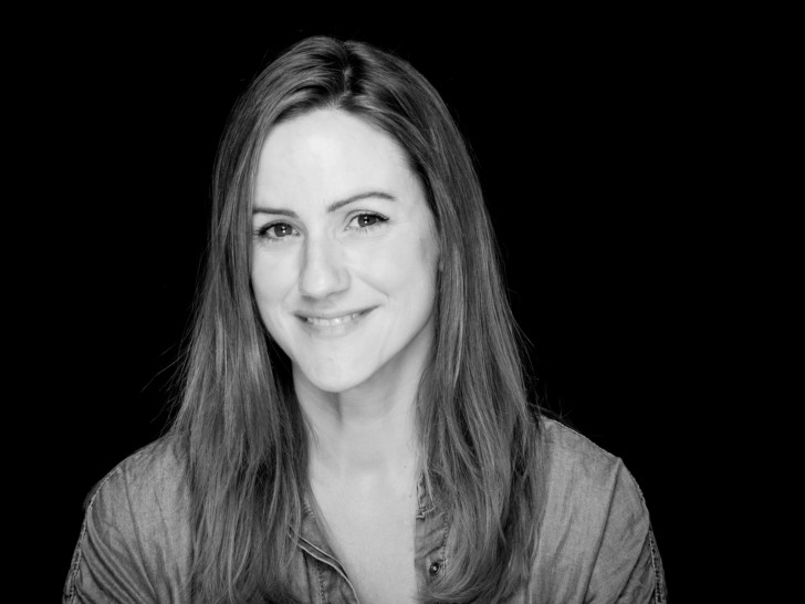 Joanna is photographed in black and white in front of a black background. She has medium length brunette hair.