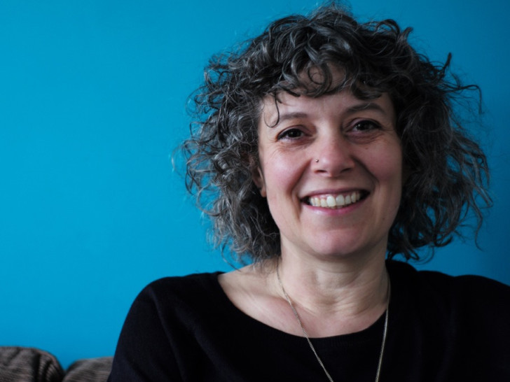 Julia smiles at the camera in front of a blue wall, she has short grey curly hair.