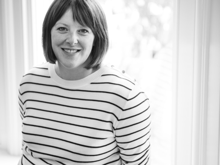 Kat is photographed in Black and White stood in front of a window, smiling at the camera. She has Short hair with a fringe and is wearing a striped long sleeve top.