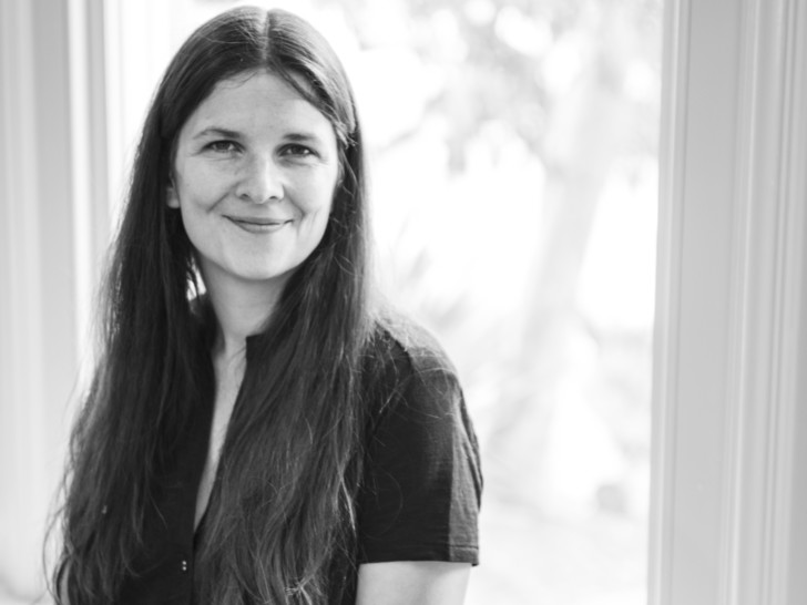 Angharad is photographed in Black and White smiling at the camera in front of a window. She wears a short sleeved blouse and has long, dark hair.