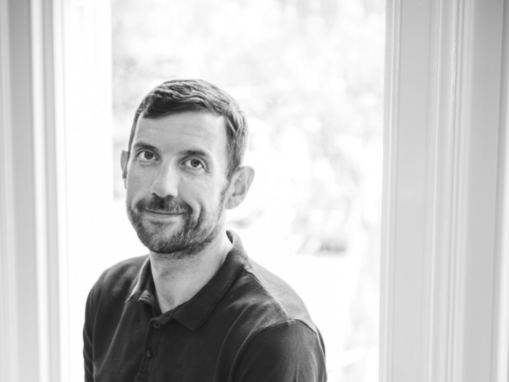 Patrick is smiling a the camera in this black and white photo. He has short dark hair and wears a black t shirt.