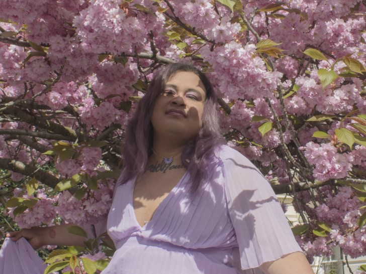 Arthur Britney Joestar is looking at the camera, they are wearing a lilac dress and are standing underneath a tree that is blossoming.