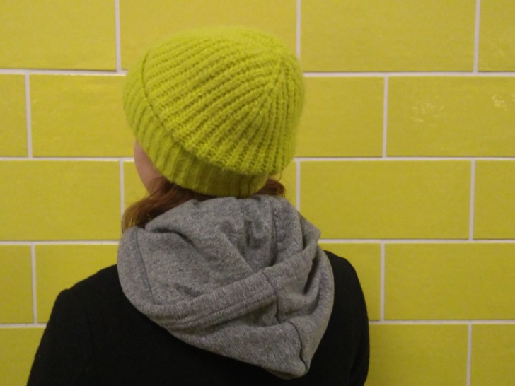 Emily is photographed with their back to the camera, they are wearing a lime green knitted hat and are stood in front of a lime green wall.