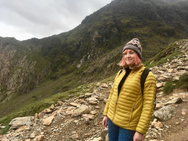 Lucie is wearing a yellow puffer jacket and a bobble hat, she is photographed in front of a mountain looking away from the camera and has short pink hair.
