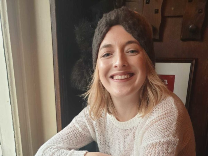 Rhyannon is photographed smiling at the camera sat at a wooden table, she has short blonde hair and is wearing a grey knitted headband.