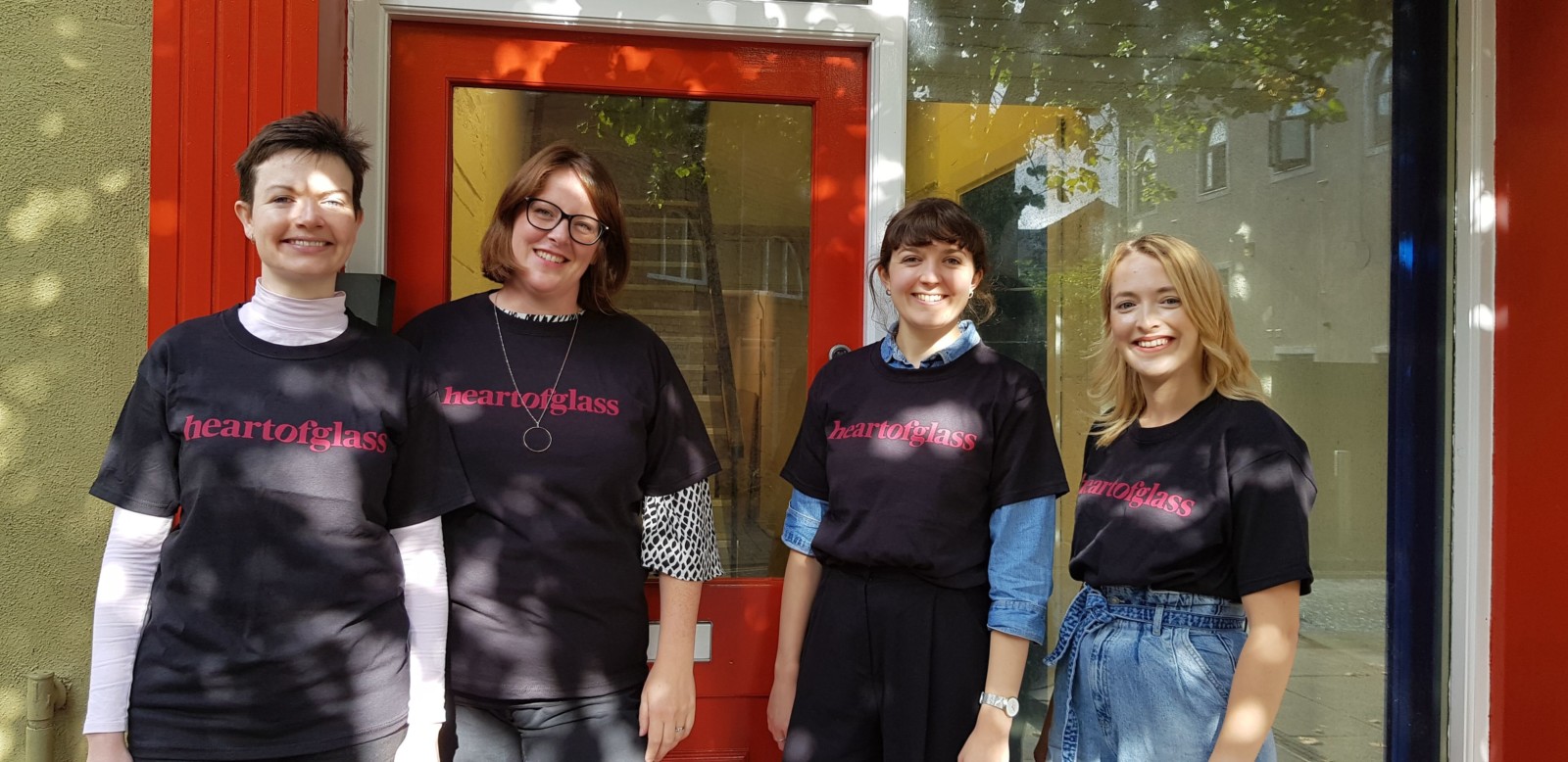 Four women stand in front of a building with a red door and woodwork and a large window. They are all wearing black tshirts with the red Heart of Glass logo printed on to them. This new logo is the words heart of glass across one horizontal line without spaces.